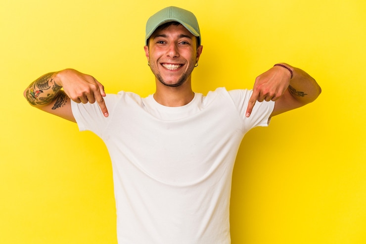 Foto jovem homem caucasiano com tatuagens isoladas em um fundo amarelo aponta para baixo com os dedos, sentimento positivo.