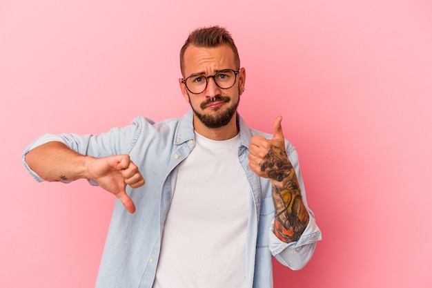 Jovem homem caucasiano com tatuagens isoladas em um fundo rosa mostrando os polegares para cima e para baixo, é difícil escolher o conceito