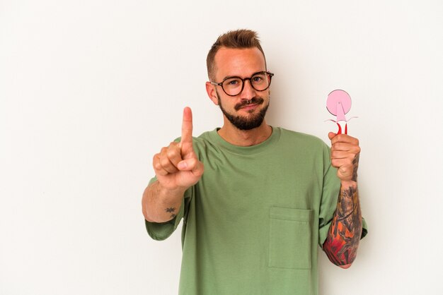 Jovem homem caucasiano com tatuagens segurando pirulito isolado no fundo branco, mostrando o número um com o dedo.