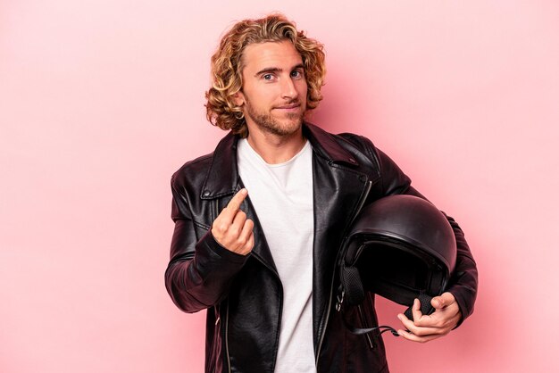 Jovem homem caucasiano com um capacete de moto isolado no fundo rosa, apontando com o dedo para você como se estivesse convidando para se aproximar.