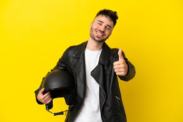 Foto jovem homem caucasiano com um capacete de motociclista isolado em um fundo amarelo com polegar para cima porque algo de bom aconteceu