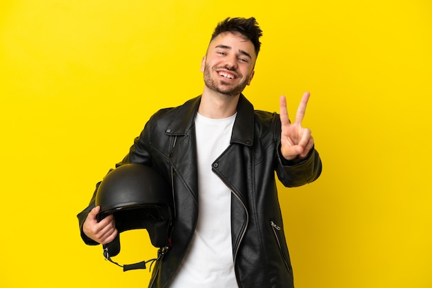 Jovem homem caucasiano com um capacete de motociclista isolado em um fundo amarelo, sorrindo e mostrando sinal de vitória