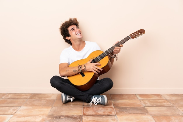 Jovem homem caucasiano com uma guitarra, sentada no chão, olhando para cima enquanto sorrindo