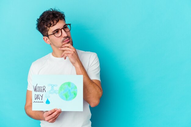 Jovem homem caucasiano comemorando o dia mundial da água isolado, olhando de soslaio com expressão duvidosa e cética.