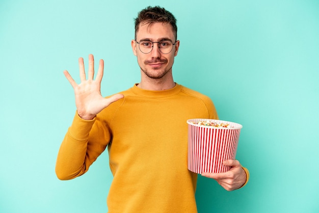 Jovem homem caucasiano comendo pipoca isolada em fundo azul, sorrindo alegre mostrando o número cinco com os dedos.