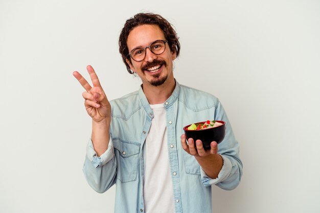 Jovem homem caucasiano comendo um ramen isolado no branco, alegre e despreocupado, mostrando um símbolo da paz com os dedos.