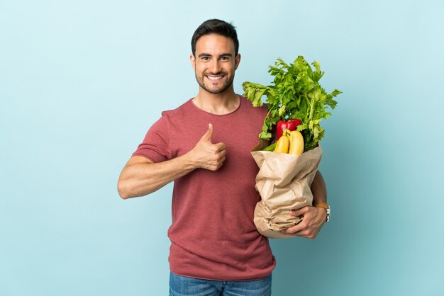 Jovem homem caucasiano comprando alguns vegetais isolados em azul e fazendo um gesto de polegar para cima