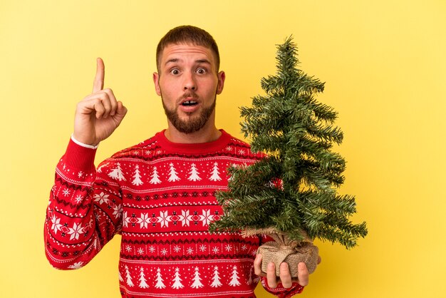 Jovem homem caucasiano comprando arvore de Natal isolada em fundo amarelo, tendo uma ideia, o conceito de inspiração.