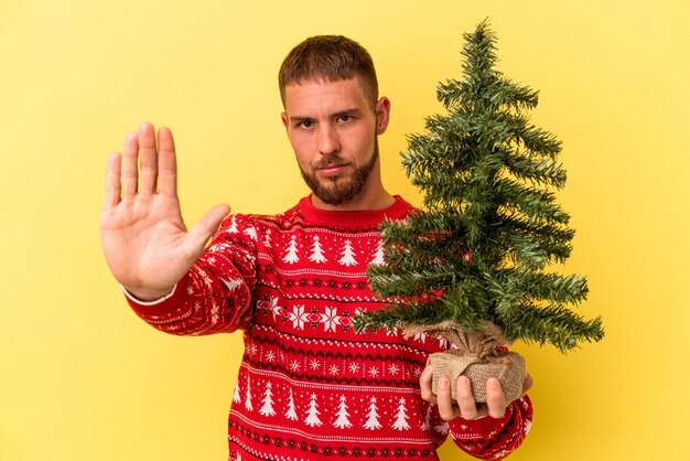 Jovem homem caucasiano comprando arvore de Natal isolada em um fundo amarelo, de pé com a mão estendida, mostrando o sinal de pare, impedindo-o.