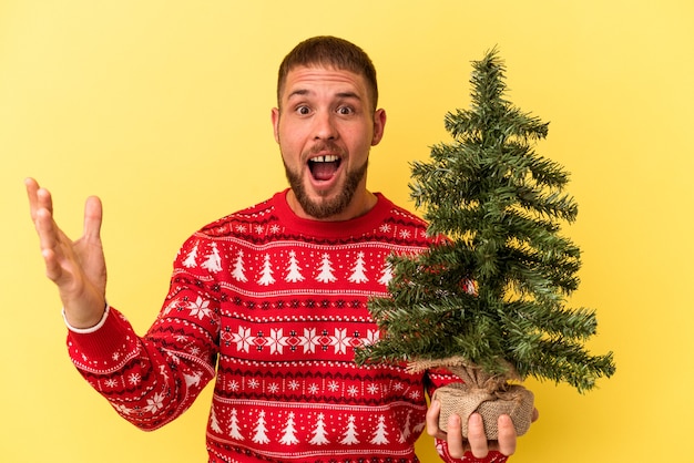 Jovem homem caucasiano comprando arvore de Natal isolada no fundo amarelo, recebendo uma agradável surpresa, animado e levantando as mãos.