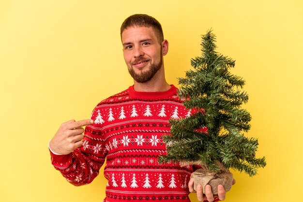 Jovem homem caucasiano comprando uma pequena árvore para o Natal isolada em um fundo amarelo pessoa apontando com a mão para um espaço de cópia de camisa, orgulhoso e confiante