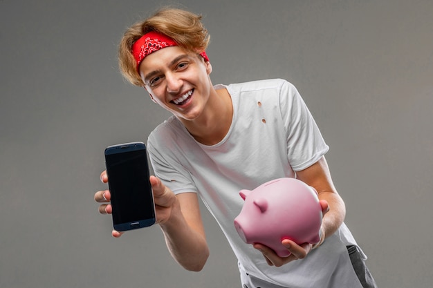 Jovem homem caucasiano em óculos de sol vermelhos, t-shirt branca com caixa de dinheiro porco rosa na parede cinza