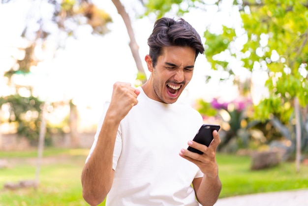 Jovem homem caucasiano em um parque usando telefone celular e fazendo gesto de vitória