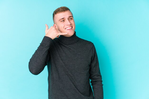 Jovem homem caucasiano em uma parede azul, mostrando um gesto de chamada de telefone móvel com os dedos.