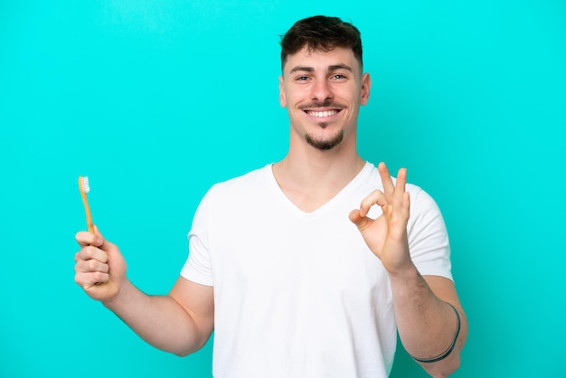 Jovem homem caucasiano escovando os dentes isolados em fundo azul, mostrando sinal de ok com os dedos