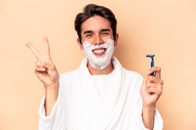 Jovem homem caucasiano fazendo a barba isolada em fundo bege alegre e despreocupado, mostrando um símbolo de paz com os dedos.