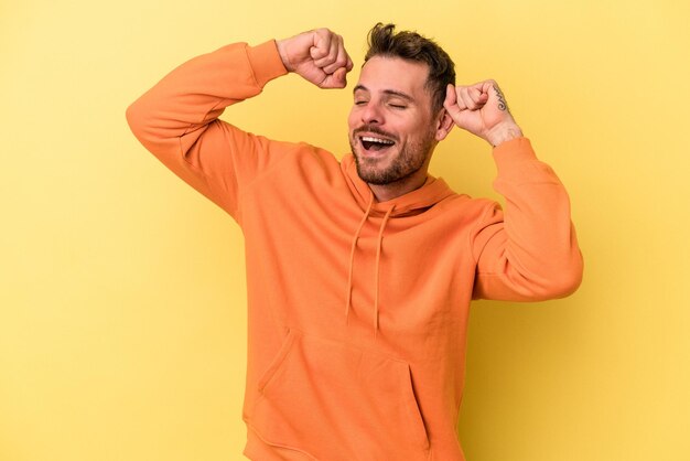 Jovem homem caucasiano isolado em fundo amarelo comemorando um dia especial, pula e levanta os braços com energia.