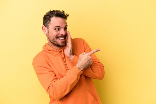 Jovem homem caucasiano isolado em fundo amarelo dizendo uma fofoca, apontando para o lado relatando algo.