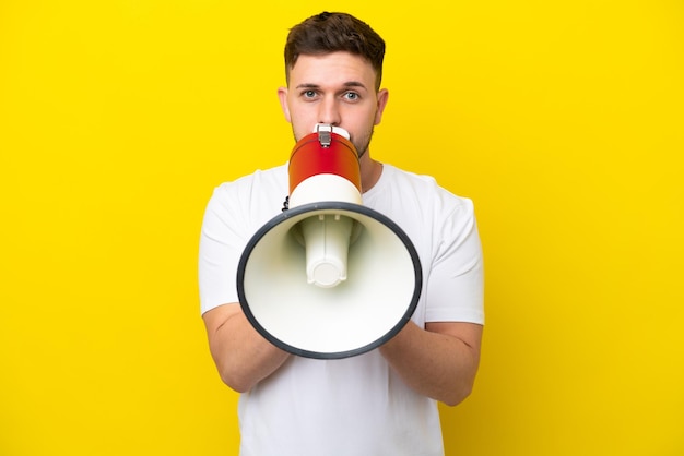 Jovem homem caucasiano isolado em fundo amarelo gritando através de um megafone para anunciar algo