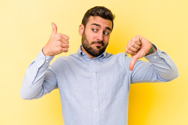 Jovem homem caucasiano isolado em fundo amarelo, mostrando os polegares para cima e os polegares para baixo, difícil escolher o conceito