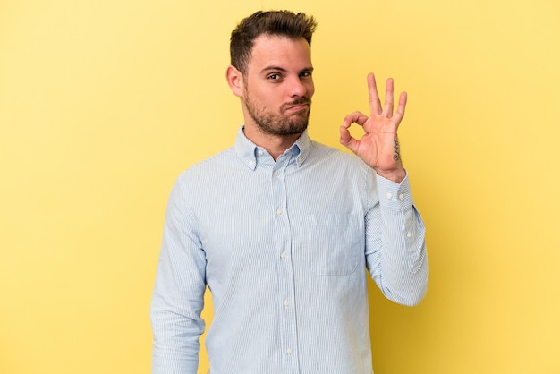 Foto jovem homem caucasiano isolado em fundo amarelo pisca um olho e mantém um gesto bem com a mão.