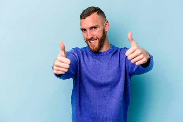 Jovem homem caucasiano isolado em fundo azul, levantando os dois polegares, sorrindo e confiante.