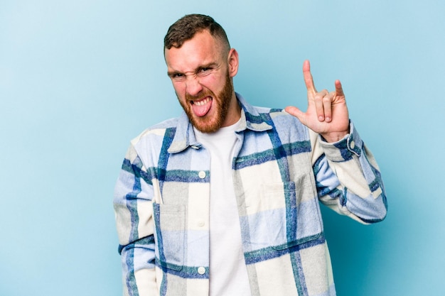 Jovem homem caucasiano isolado em fundo azul, mostrando o gesto de rock com os dedos