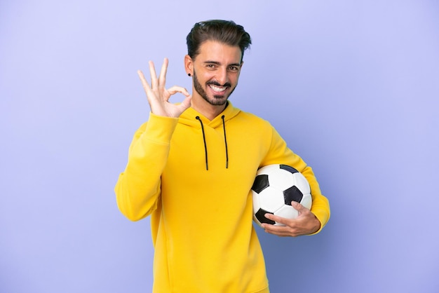 Jovem homem caucasiano isolado em fundo roxo com bola de futebol e fazendo sinal de OK
