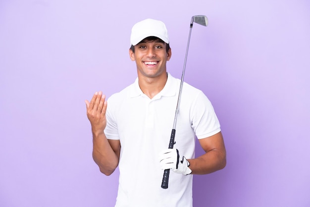 Jovem homem caucasiano isolado em fundo roxo jogando golfe e fazendo gesto de vinda