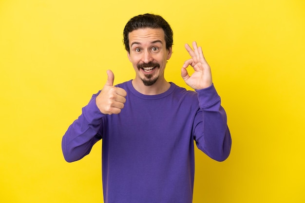 Jovem homem caucasiano isolado em um fundo amarelo mostrando sinal de ok e gesto de polegar para cima