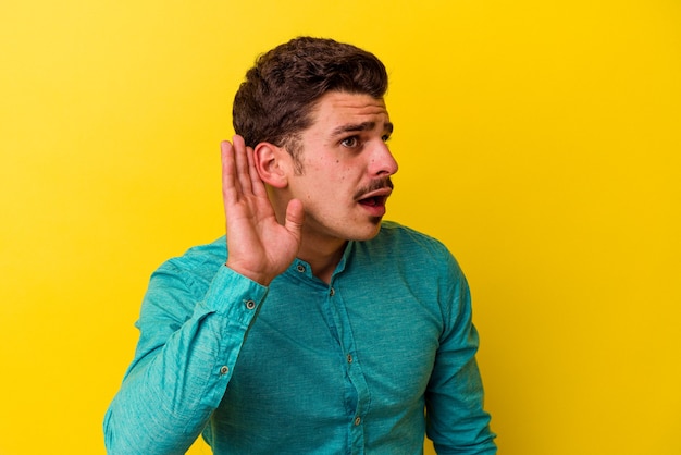 Jovem homem caucasiano isolado em um fundo amarelo, tentando ouvir uma fofoca.