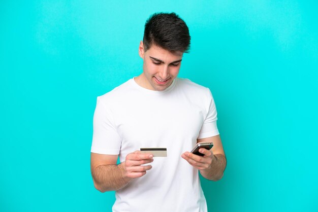 Jovem homem caucasiano isolado em um fundo azul comprando com o celular com um cartão de crédito