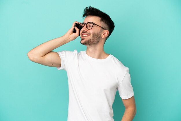 Jovem homem caucasiano isolado em um fundo azul, conversando com alguém ao telefone celular