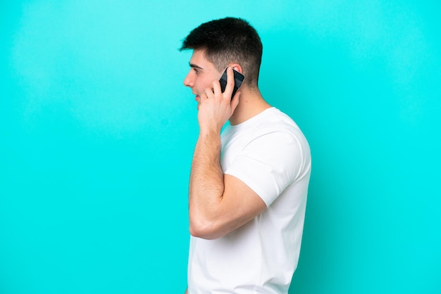 Jovem homem caucasiano isolado em um fundo azul, conversando com alguém ao telefone celular.