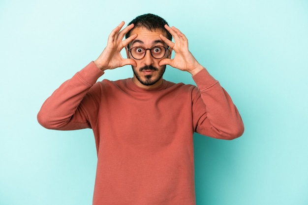 Jovem homem caucasiano isolado em um fundo azul, mantendo os olhos abertos para encontrar uma oportunidade de sucesso.