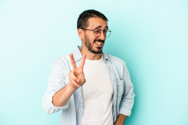 Jovem homem caucasiano isolado em um fundo azul, mostrando o número dois com os dedos.