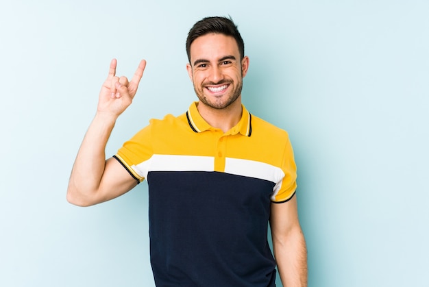 Jovem homem caucasiano isolado em um fundo azul, mostrando um gesto de chifres como um conceito de revolução.