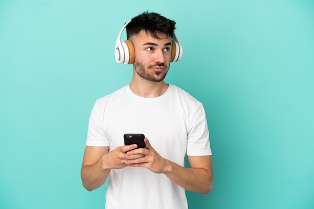 Jovem homem caucasiano isolado em um fundo azul ouvindo música com um celular e pensando
