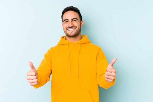 Jovem homem caucasiano isolado em um fundo azul, sorrindo e levantando o polegar