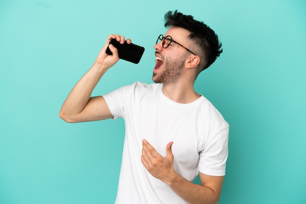 Jovem homem caucasiano isolado em um fundo azul usando telefone celular e cantando