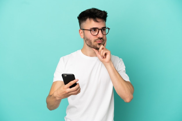 Jovem homem caucasiano isolado em um fundo azul usando telefone celular e pensando