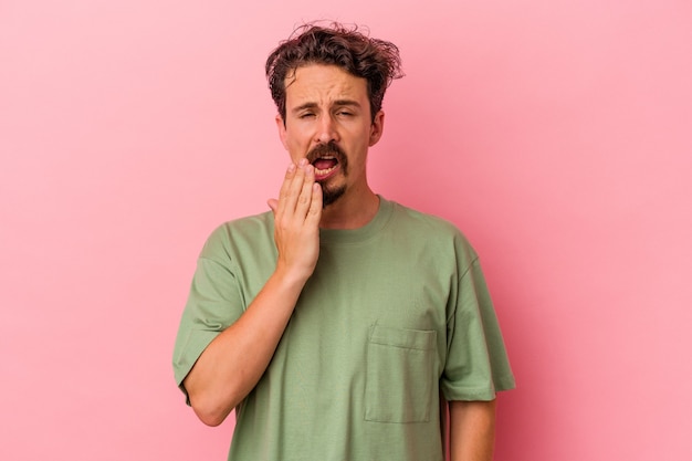 Jovem homem caucasiano isolado em um fundo rosa bocejando, mostrando um gesto cansado, cobrindo a boca com a mão.