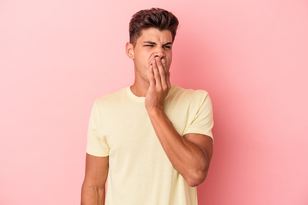 Jovem homem caucasiano isolado em um fundo rosa bocejando, mostrando um gesto cansado, cobrindo a boca com a mão.