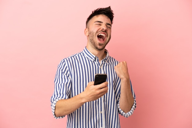 Jovem homem caucasiano isolado em um fundo rosa com o telefone em posição de vitória