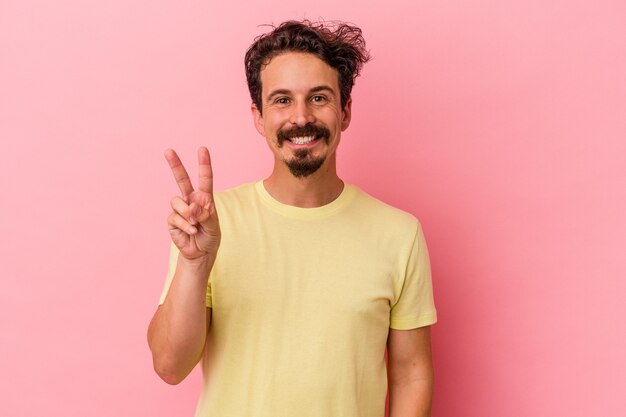 Jovem homem caucasiano isolado em um fundo rosa, mostrando sinal de vitória e sorrindo amplamente.