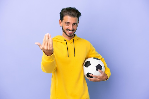 Jovem homem caucasiano isolado em um fundo roxo com uma bola de futebol e fazendo o gesto de aproximar-se