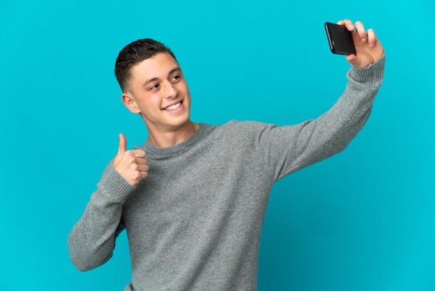 Jovem homem caucasiano isolado na parede azul fazendo uma selfie com o celular