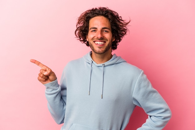 Jovem homem caucasiano isolado na rosa bakcground sorrindo alegremente, apontando com o dedo indicador afastado.