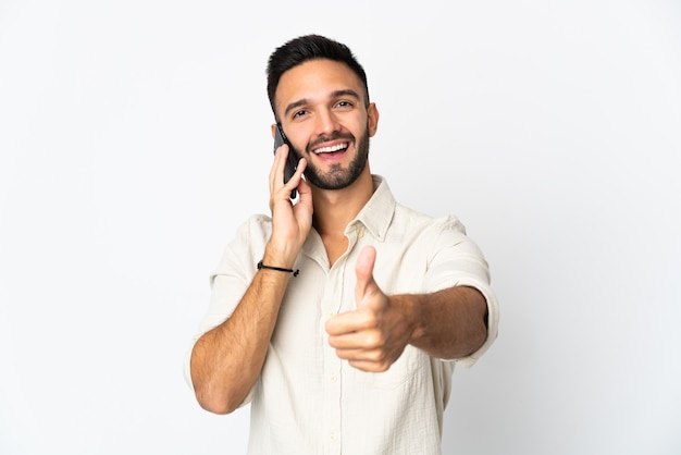 Jovem homem caucasiano isolado na superfície branca, conversando com o celular enquanto faz o polegar para cima