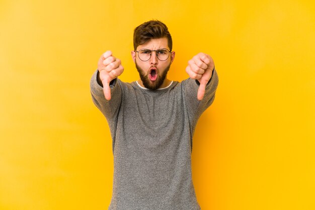 Jovem homem caucasiano isolado no espaço amarelo, mostrando o polegar para baixo e expressando antipatia.